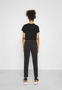 Donna con capelli ricci che indossa una maglietta nera, jeans grigi scuri a vita alta, calzini neri e sneakers bianche con dettagli gialli e verdi.
