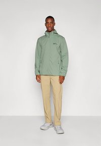 Uomo con giacca leggera verde chiaro con zip, pantaloni beige e sneakers bianche, in piedi contro uno sfondo grigio chiaro uniforme.
