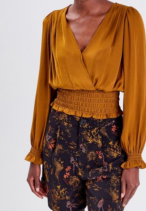 Femme portant un chemisier cache-cœur à manches longues moutarde avec taille froncée et short taille haute noir à motif floral.
