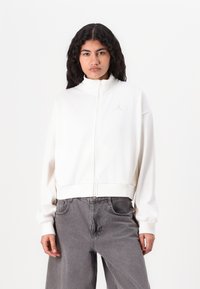 Hvid zip-up jakke med høj krave og ribbede manchetter, prydet med et diskret guld-logo på venstre bryst. Båret med grå vide bukser.