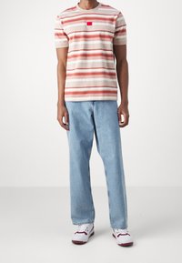 Stribet kortærmet t-shirt i beige, rød og lys rosa med et lille rødt logo-patch, parret med lyseblå afslappede jeans og hvide sneakers.