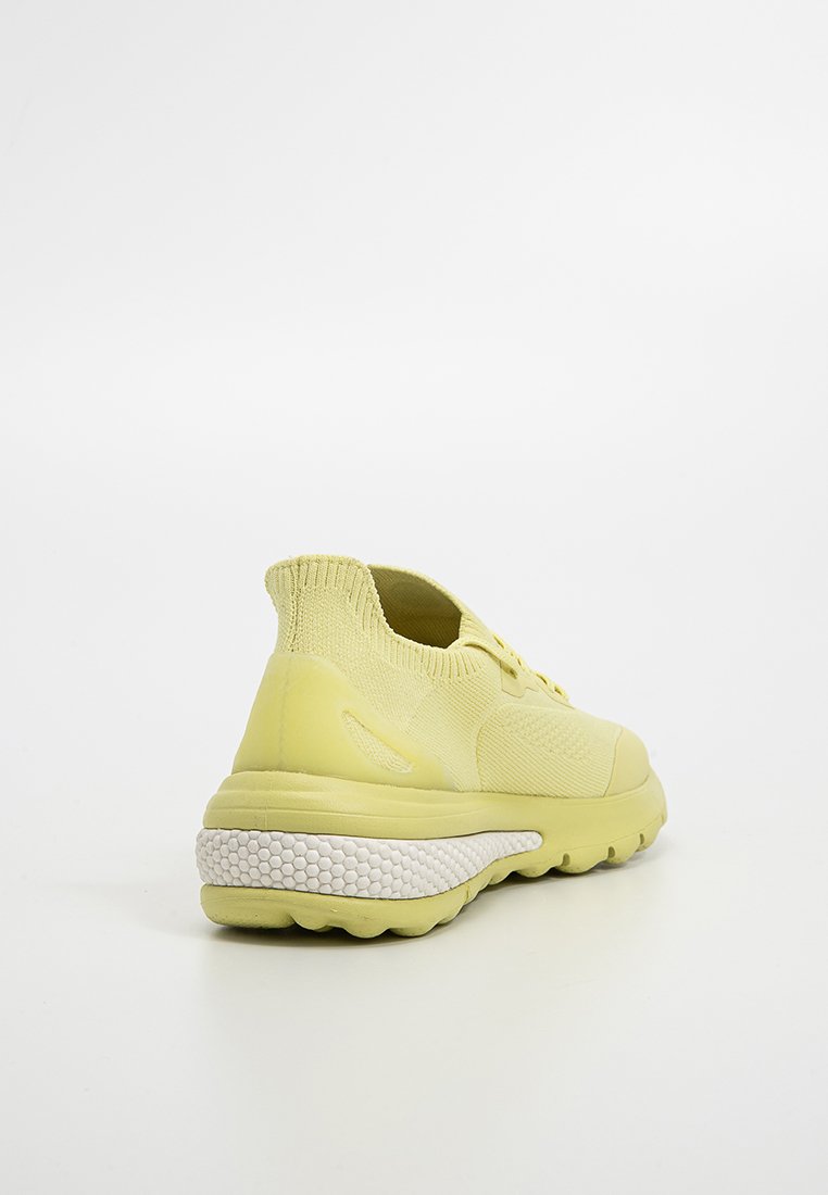 Zapatilla amarilla con parte superior de punto, collar de perfil bajo y suela blanca texturizada. Forma redondeada con soporte acolchado en el talón.