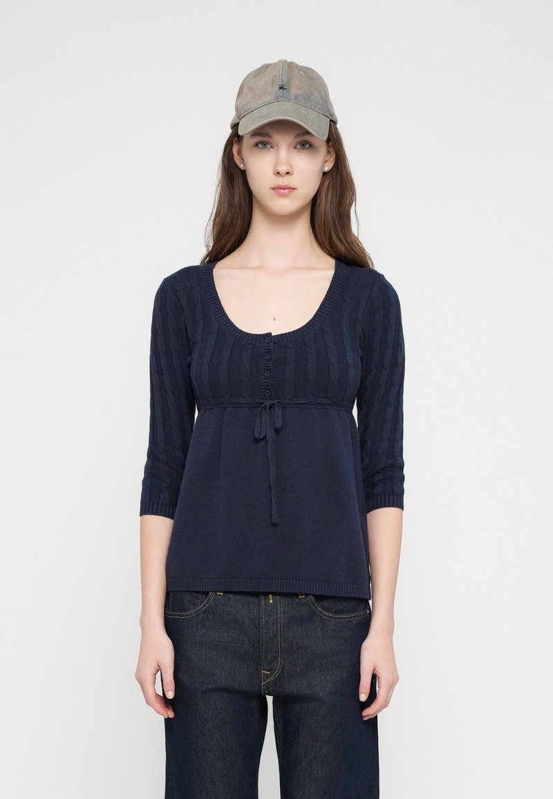 Μπλε πλεκτό τοπ με υφασμάτινες πιέτες, στρογγυλή λαιμόκοψη, κουμπιά στο μπροστινό μέρος και λεπτομέρεια δέσιμο στη μέση. Συνδυασμένο με σκούρο τζιν παντελόνι και καπέλο.