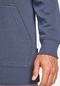 Felpa blu scuro con orlo e polsini a costine. Presenta una tasca frontale con apertura diagonale. Il materiale sembra morbido e testurizzato.