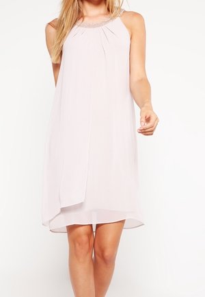 Femme portant une robe rose clair sans manches, longueur genou, avec un col orné de perles, debout devant un fond blanc uni.