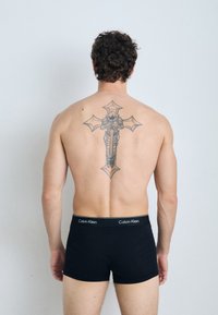 Zwarte katoenen boxershorts met een Calvin Klein-logo op de tailleband, gedragen door een persoon met krullend haar en een gedetailleerde rugtattoo.