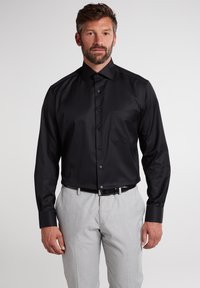 Schwarz langärmliges Hemd mit glatter Textur, mit Brusttasche und Knopfverschluss. Kombiniert mit hellgrauen Hosen und einem schwarzen Gürtel.