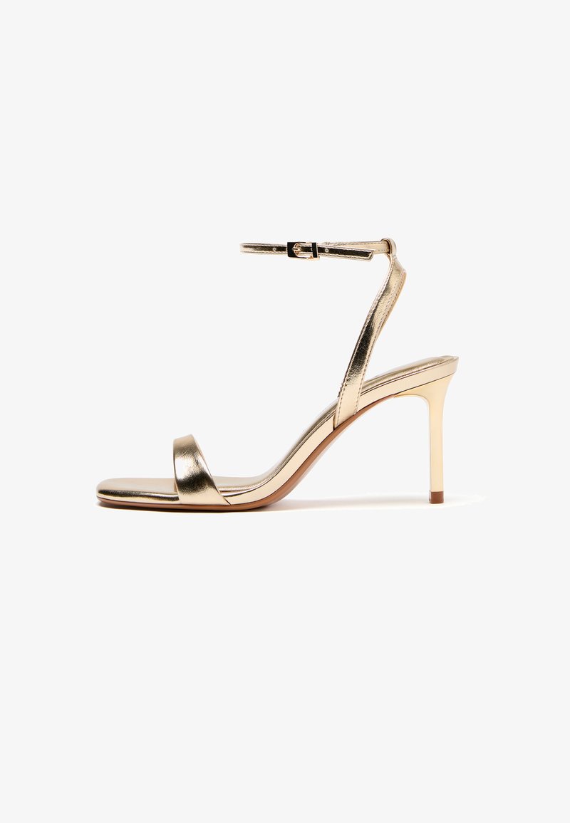 Zlatá dámska sandála na vysokom podpätku s opaskom okolo členka, otvorenou špičkou, tenkým ihľovým podpätkom a malou prackou na bielom pozadí.