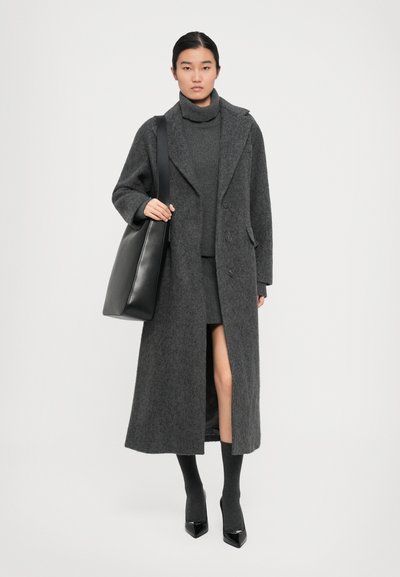 Long manteau en laine gris avec un col à revers, associé à un pull-over gris et une jupe. Un sac fourre-tout noir et des escarpins noirs brillants complètent le look.