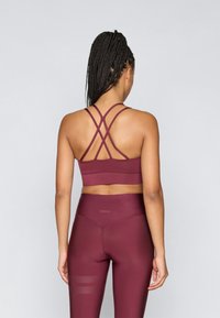 Μπορντό αθλητικό σουτιέν με σταυρωτές τιράντες και ταιριαστά ψηλόμεσα κολάν. Το ύφασμα φαίνεται λείο με διακριτικά σχέδια.