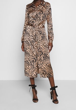 Vrouw draagt een midi-jurk met lange mouwen met een beige en zwarte zebraprint, een riem en zwarte sandalen met hoge hakken en strikjes bij de enkels.