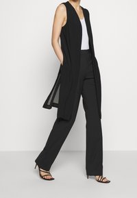 Svart ärmlös lång väst med sidofickor, tillsammans med svarta vida byxor och sandaler med klack och remmar, med en jämn tygtextur.