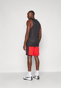 Svart mesh tank top sammen med røde idrettsshorts med sidestriper. Personen har på seg hvite og svarte sportssko med hvite sokker.