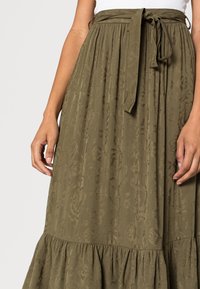 Mains sur les côtés, portant une jupe vert olive avec un motif jacquard floral et une taille nouée sur un haut blanc.