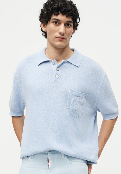 Νέος άνδρας με σγουρά σκούρα μαλλιά που φοράει φωτεινό γαλάζιο κοντομάνικο πλεκτό πόλο με κουμπωτό γιακά και υφασμένη τσέπη.