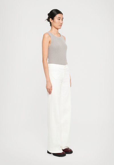 Femme vue de profil portant un haut sans manches gris, un pantalon large taille haute blanc et des chaussures plates sombres, sur fond uni.