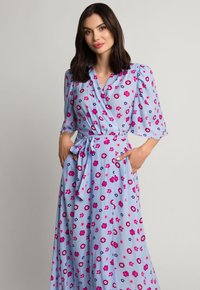 Jasnoniebieska sukienka w stylu wrap z krótkimi bufkami, paskiem w talii oraz kwiatowym wzorem w kolorze różowym, fioletowym i białym. Gładka tekstura materiału.