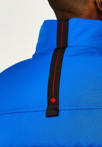Blaue Jacke mit hohem Kragen, ausgestattet mit einem schwarzen Riemen mit roter Naht und einem roten Ahornblatt-Emblem auf der Rückseite. Glatte Stofftextur.