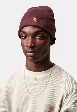 Hombre con un gorro de punto color burdeos con un logo amarillo, suéter texturizado color crema con un parche y collar de cadena plateada, frente a un fondo gris liso.