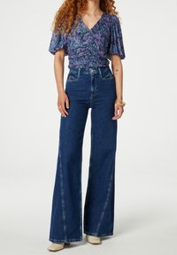 Jeans in denim blu a vita alta e gamba larga con cuciture visibili e tasche frontali, abbinati a un top viola fantasia con maniche a sbuffo.