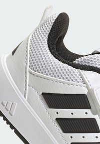 Bijela atletska cipela s gornjim dijelom od teksturiranog tkanog materijala, crnim mrežastim ovratnikom, gumeni potplat i tri crne pruge sa strane.