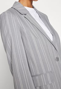 Szara marynarka w cienkie prążki z klapą z wcięciem, zapięciem na jeden guzik i przednią kieszenią. Tkanina ma gładką strukturę z cienkimi liniami.