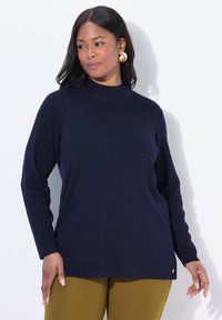 Frau trägt ein navyblaues langärmliges Oberteil und senfgelbe Hose, steht vor einem weißen Hintergrund mit goldenen Ohrringen und einem neutralen Gesichtsausdruck.