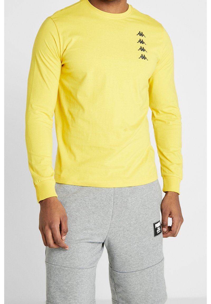 Mann trägt ein gelbes Langarmhemd mit kleinen schwarzen Logos auf der Brust und hellgraue Jogginghosen mit einem schwarzen Patch auf dem Oberschenkel.