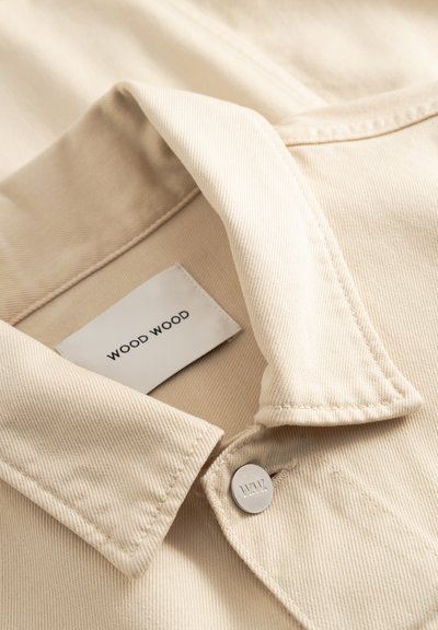 Beige denim jacket collar with visible brand label "WOOD WOOD" and a metal button engraved with "W.W." on stitched fabric.