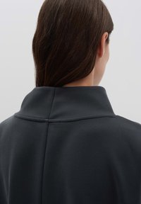 Donna con capelli marrone scuro, che indossa un indumento scuro a colletto alto, vista da dietro su uno sfondo chiaro e uniforme.