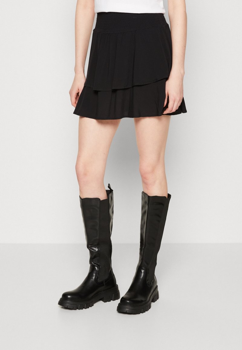 Femme portant une mini-jupe noire à volants et des bottes en cuir noires montantes jusqu'aux genoux, debout sur un fond uni.