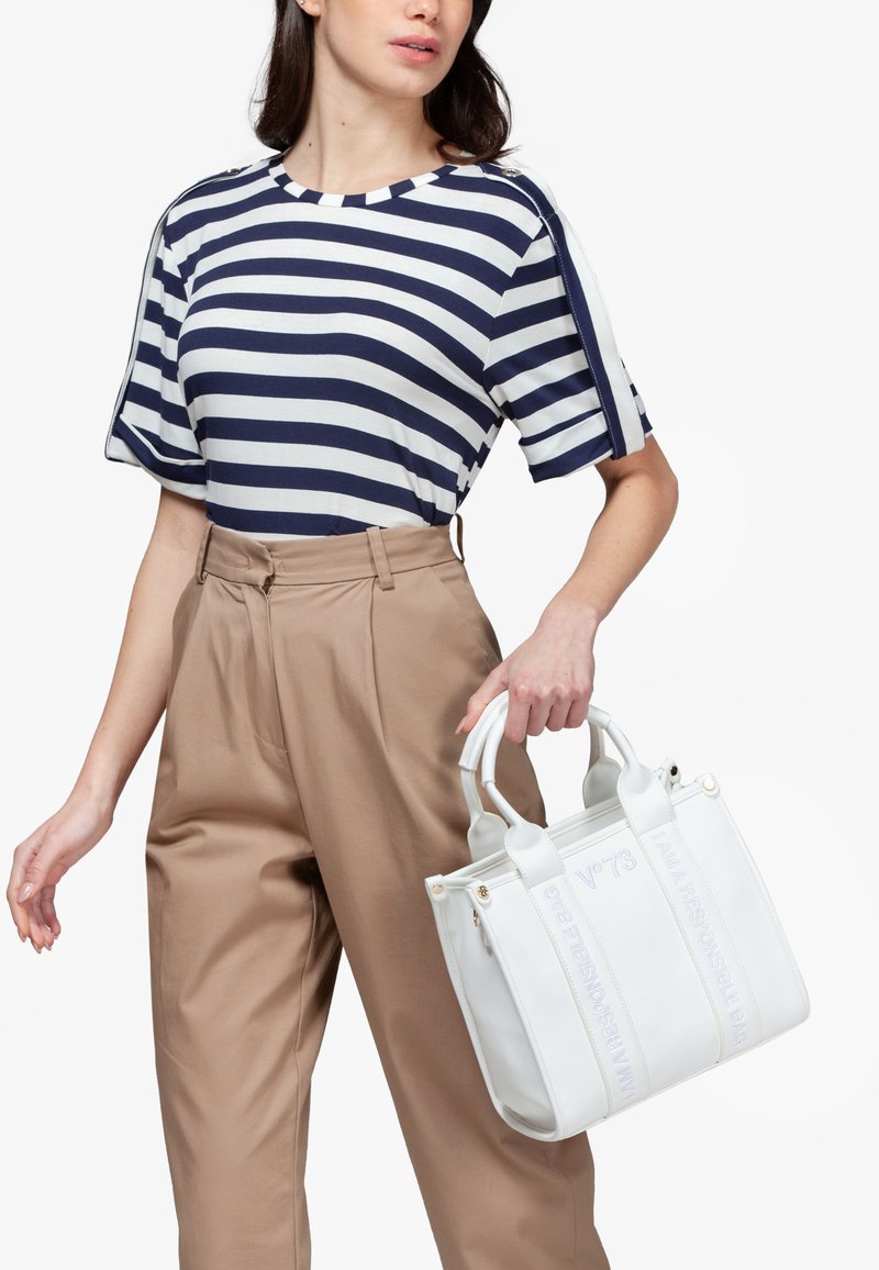 Borsa bianca con un design strutturato, dotata di due manici superiori e testo in rilievo. Abbinata a una camicia a righe blu navy e bianca e pantaloni beige.