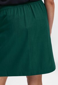 Jupe en tissu vert avec taille élastique, longueur genoux, texture lisse, détail de couture unique et un design simple, sans embellissement.