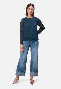 Marineblaue Bluse mit Falten an der Vorderseite und transparenten Ärmeln; kombiniert mit weit geschnittenen hellen Jeans und silbernen spitzen High Heels.