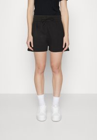 Pantaloni corti neri in cotone con elastico in vita e cordino, dotati di una vestibilità rilassata e cuciture laterali, abbinati a scarpe da ginnastica bianche e calze.