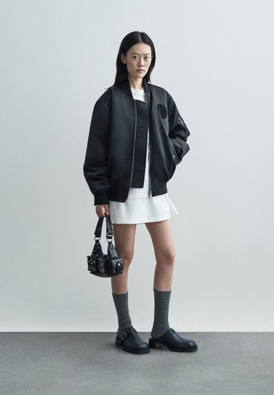 Chaqueta bomber negra con un parche redondo, vestido blanco, calcetas gris hasta la rodilla y zapatos negros gruesos. Pequeña bolsa negra con tachuelas.