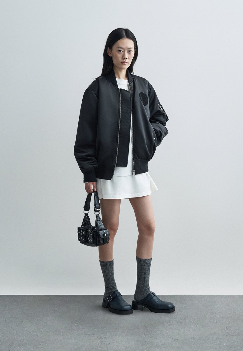 Schwarze Bomberjacke mit rundem Aufnäher, weißes Kleid, graue knielange Socken und klobige schwarze Schuhe. Kleine schwarze Tasche mit Nieten.