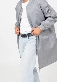 Homme portant un jean bleu clair, un t-shirt blanc et une longue veste bomber grise ajustant une ceinture noire avec une boucle argentée.