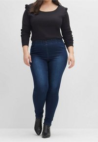 Svart långärmad topp med volangdetaljer, ihop med mörkblå, stretchiga jeans i denim med elastisk midja och avsmalnande ben.