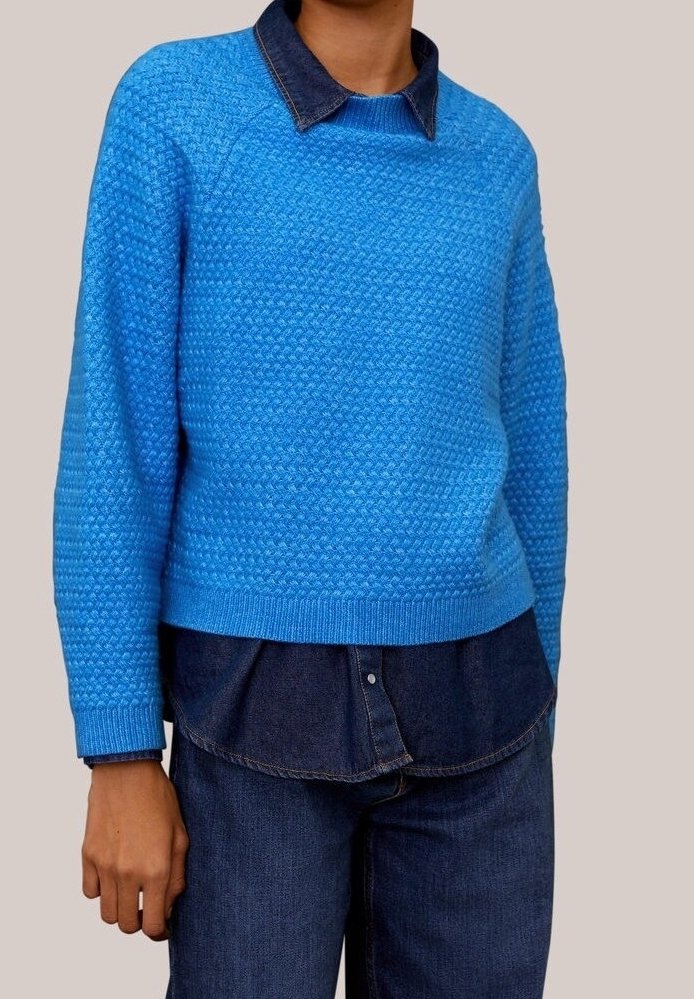 Personne portant un pull tricoté texturé bleu vif par-dessus une chemise en denim bleu foncé et un jean bleu foncé, debout devant un fond uni.