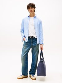 Camisa de botones azul clara sobre una camiseta blanca, combinada con jeans azules holgados y zapatillas beige. Sosteniendo una bolsa de mano azul marino.
