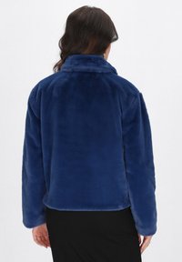 Donna con capelli scuri che indossa un giaccone in pelliccia sintetica blu royal e una gonna nera, vista di spalle su uno sfondo bianco.