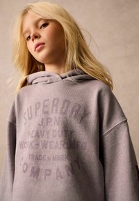 Felpa grigia oversize con una grande stampa testuale in grigio scuro. Il tessuto appare morbido con una superficie liscia e una cappuccio regolabile con lacci.