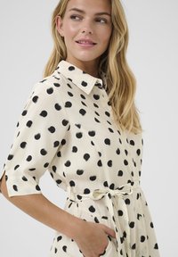 Abito camicia color crema con grandi pois neri, maniche corte, colletto, chiusura a bottoni sul davanti e cintura annodata in vita. Tessuto dalla texture liscia.