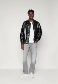 Hombre con dreadlocks cortos que lleva una chaqueta bomber negra brillante, camiseta blanca, jeans gris claro y zapatillas blancas, de pie con calma.