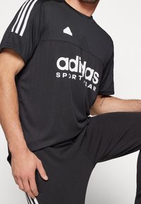 Černé sportovní tričko s krátkými rukávy, se bílým logem "adidas SPORTWEAR" a třemi bílými pruhy na ramenou.