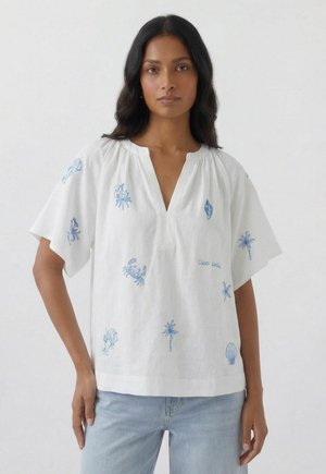 Frau trägt ein weißes, lockeres Kurzarmshirt mit blau bestickten Meeresbewohnern und Palmen, kombiniert mit hellblauen Jeans.