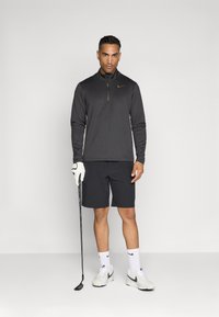 Crna zip-up majica dugih rukava s žutim logom, u kombinaciji s crnim šortsevima. Bijela golf rukavica i cipele, drži golf štap. Jednostavna pozadina.