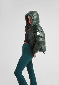 Kort grøn puff jakke med hætte, der har en glat finish og et hexagonalt logo-patch på venstre arm. Sæt sammen med teal leggings.