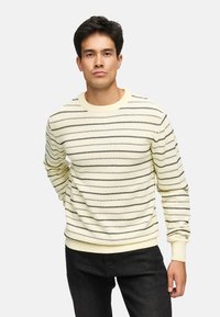 Lys gul strikket sweater med sorte vandrette striber, rund hals og ribstrikkede cuffs og kant. Modellen har mørkegrå bukser på.